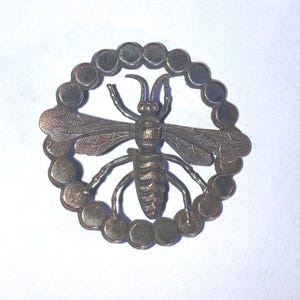 May include: A circular metal brooch featuring a central bee motif with detailed wings and body. The bee is enclosed within a ring composed of small, connected metallic spheres. The entire piece has a dark, tarnished finish with visible texture.