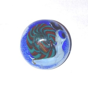May include: A round glass marble featuring a snail motif. The snail has a light blue body and a spiraled shell with swirling patterns of dark red, teal, and brown. The background of the marble is a deep blue with a textured, speckled appearance.