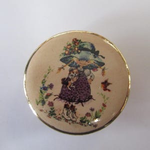 May include: A round, cream-colored trinket box with a gold-tone metal rim. The lid features a printed illustration of a figure wearing a wide-brimmed blue hat and a purple dress, holding a small dog, surrounded by colorful floral accents and a bird.