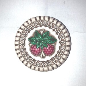 May include: A round ceramic button featuring a central design of two pink raspberries with green leaves. The border is decorated with a white and brown scalloped pattern and a series of small vertical lines around the outer edge of the button.