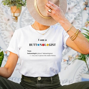 May include: White t-shirt with the text "I am a BUTTONOLOGIST" and a definition of the word. The word "BUTTONOLOGIST" has colorful buttons replacing the "O"s.