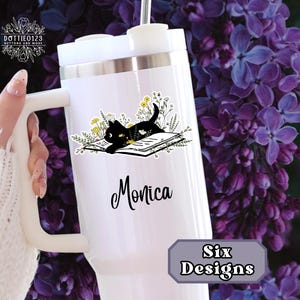 May include: White tumbler with a black cat illustration on a book, surrounded by yellow flowers. The name "Monica" is written in black script. The tumbler has a white handle and a silver rim. The text "Six Designs" is in a gray rectangle.