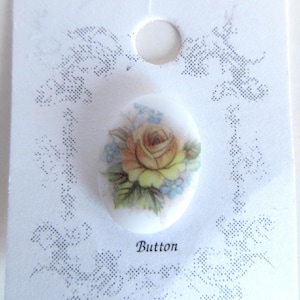 May include: An oval-shaped button with a floral design featuring a yellow rose and blue accents. The button is attached to a white card with the word "Button" printed on it. The card has a decorative dotted border.