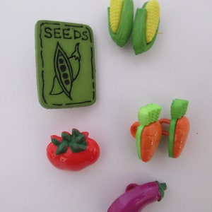 May include: Assortment of colorful plastic buttons shaped like vegetables, including a green pea pod, a red tomato, a purple eggplant, two orange carrots, and two corn cobs. The buttons have a green "SEEDS" label.