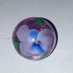 May include: A spherical glass marble featuring a translucent purple base with an internal floral design. The flower has light purple and blue petals with a small yellow center, accented by small green leaf-like shapes embedded within the glass.