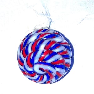 May include: A round glass button featuring a swirling, candy-like pattern of alternating red, white, and blue stripes. The surface is smooth and glossy, with the stripes radiating from the center in a continuous, coiled, and circular design.