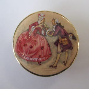 May include: A round, gold-rimmed compact featuring a painted illustration of two figures in 18th-century attire. One figure wears a voluminous pink gown with floral details, while the other wears a purple coat and breeches, both with white wigs.
