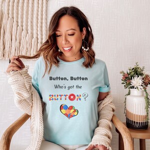 May include: A light blue t-shirt with the text "Button, Button Who's got the BUTTON?" and a heart-shaped design made of colorful buttons.