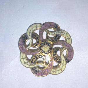 May include: A circular metal brooch featuring an intricate openwork design of interlocking curved segments. The surface is decorated with enamel in shades of cream, mauve, and black, accented with gold-toned metal edges and a central floral motif.