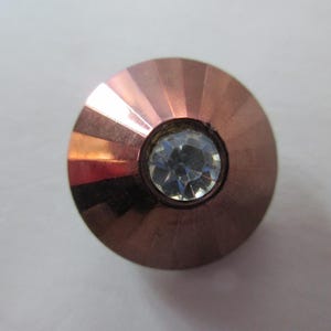 May include: A copper-colored, faceted knob with a central, clear crystal. The knob has a circular shape with a slightly domed top. The crystal is round and reflects light, adding a touch of sparkle. The copper finish has a metallic sheen.