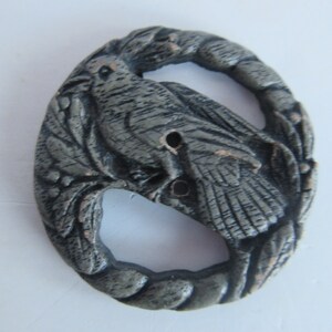 May include: A silver-toned metal button with a detailed design of a bird perched on a branch. The button has a circular shape and a hole in the center for sewing.