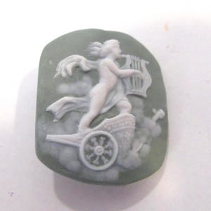 May include: A cameo in shades of green and white, featuring a classical figure playing a harp while riding a chariot. The figure is depicted in a flowing gown, with detailed relief work. The cameo has a rounded, irregular shape.