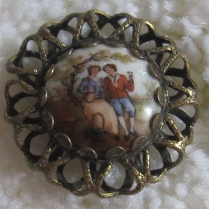 May include: A gold-toned metal button with a porcelain cameo featuring a couple in a romantic setting. The cameo is surrounded by a decorative, intricate design.