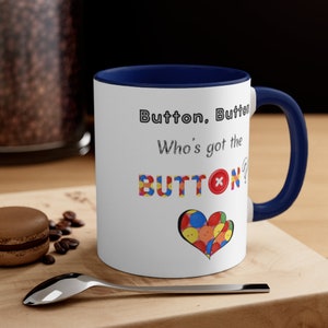 May include: White ceramic coffee mug with a blue rim and handle. The mug has a design of colorful buttons with the text "Button. Button. Who's got the BUTTON?"