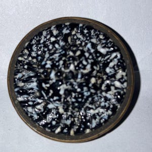 May include: A circular object with a metallic bronze-toned rim and a textured, glossy inlay featuring a dense, speckled pattern of irregular black and white shapes. The surface has a reflective, polished finish with a high-contrast appearance.