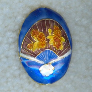 May include: A blue oval-shaped enamel pin with a gold rim. The pin features a stylized fan with brown and white stripes and yellow and orange flowers.