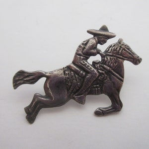 May include: A silver-toned brooch depicting a man riding a horse. The man is wearing a wide-brimmed hat and traditional clothing. The horse is in mid-gallop.