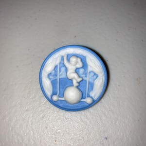 May include: A round, blue and white cameo featuring a circus performer balancing on a ball. The cameo has a detailed design of a figure, curtains, and a ball, set against a blue background.