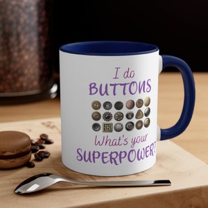May include: White ceramic coffee mug with a blue rim and handle. The mug has a purple text that reads "I do buttons" and a graphic of various buttons. The text "What's your superpower?" is also printed in purple.