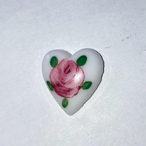 May include: A heart-shaped, white decorative item featuring a hand-painted pink rose with green leaves. The rose is centered on the heart, and the leaves are placed around the flower. The item is likely made of ceramic or glass.
