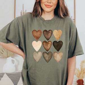 以下が含まれることがあります： オリーブグリーンのTシャツ。水彩画風の茶色のハートが9つ、3列3段の格子状に描かれています。シンプルながらも印象的なデザインです。ゆったりとしたフィット感のTシャツです。