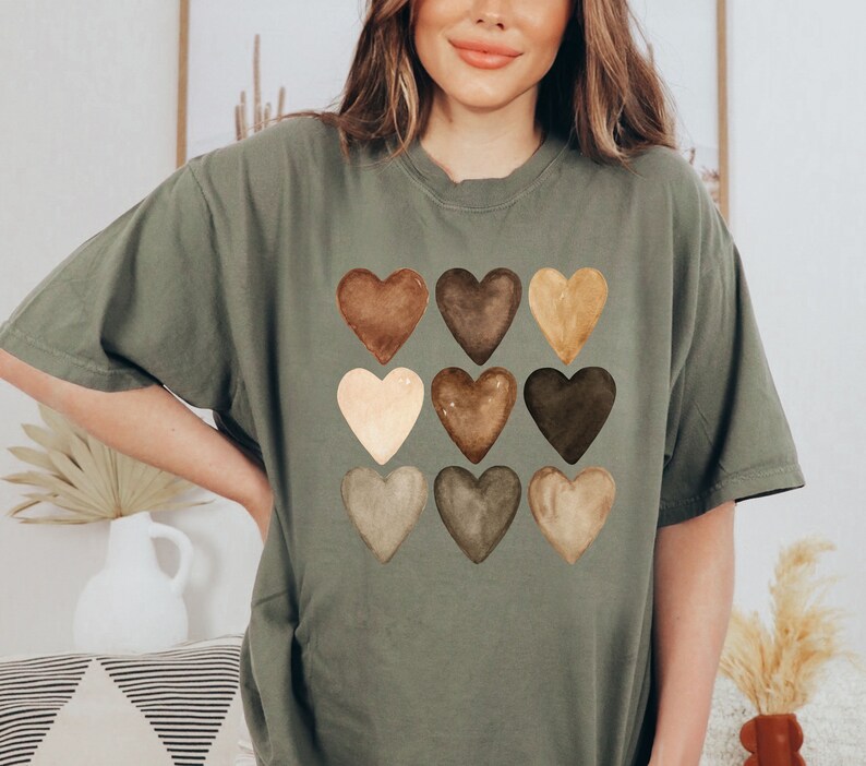 以下が含まれることがあります： オリーブグリーンのTシャツ。水彩画風の茶色のハートが9つ、3列3段の格子状に描かれています。シンプルながらも印象的なデザインです。ゆったりとしたフィット感のTシャツです。