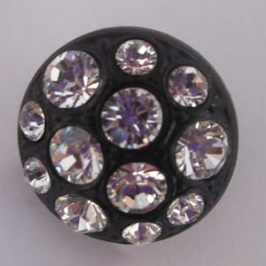May include: A black button with a round shape and a textured surface. The button is adorned with 13 clear rhinestones of varying sizes.