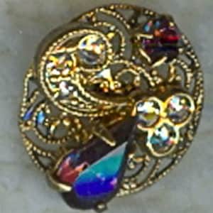 May include: A gold-toned filigree brooch with a teardrop-shaped blue and purple iridescent stone in the center. The brooch is decorated with smaller iridescent stones and has a floral design.