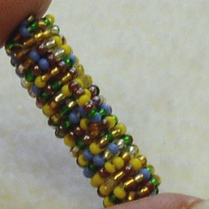 May include: A close-up of a beaded bracelet with a multi-colored pattern. The bracelet is made with small, round beads in shades of green, yellow, blue, and brown. The beads are arranged in a repeating pattern, creating a visually appealing design.