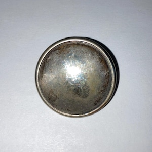 May include: A silver-toned round button with a slightly raised, domed surface. The button has a smooth, polished finish.