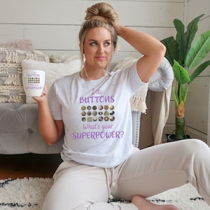 May include: A white t-shirt with the text "I do buttons, what's your superpower?" in purple. The shirt features a variety of buttons in different colors and shapes.