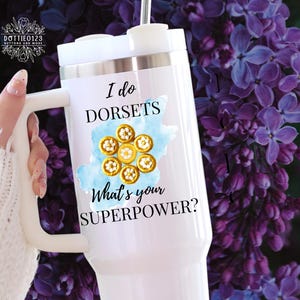 May include: White travel mug with a handle, featuring the text "I do DORSETS, What's your SUPERPOWER?" in black. The design includes a blue watercolor shape and gold-colored Dorset buttons. The mug is held against a background of purple flowers.