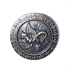 May include: A black metal button with a gold border and a detailed design of a winged creature with a serpent tail.