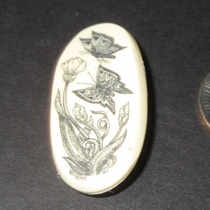 May include: A white oval-shaped pendant with a black and white engraved design of two butterflies and a flower. The pendant is signed "McClelland".