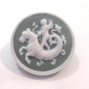 May include: A round, white and green cameo button featuring a mythological scene. A white figure with wings rides a seahorse. The button has a white border and a green background.