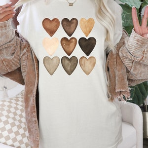 以下が含まれることがあります： クリーム色のTシャツ。9つの水彩画のハートが様々な茶色で描かれています。ハートはグリッドパターンに配置され、シンプルながらもインパクトのあるデザインです。柔らかい素材で作られており、カジュアルウェアに最適です。