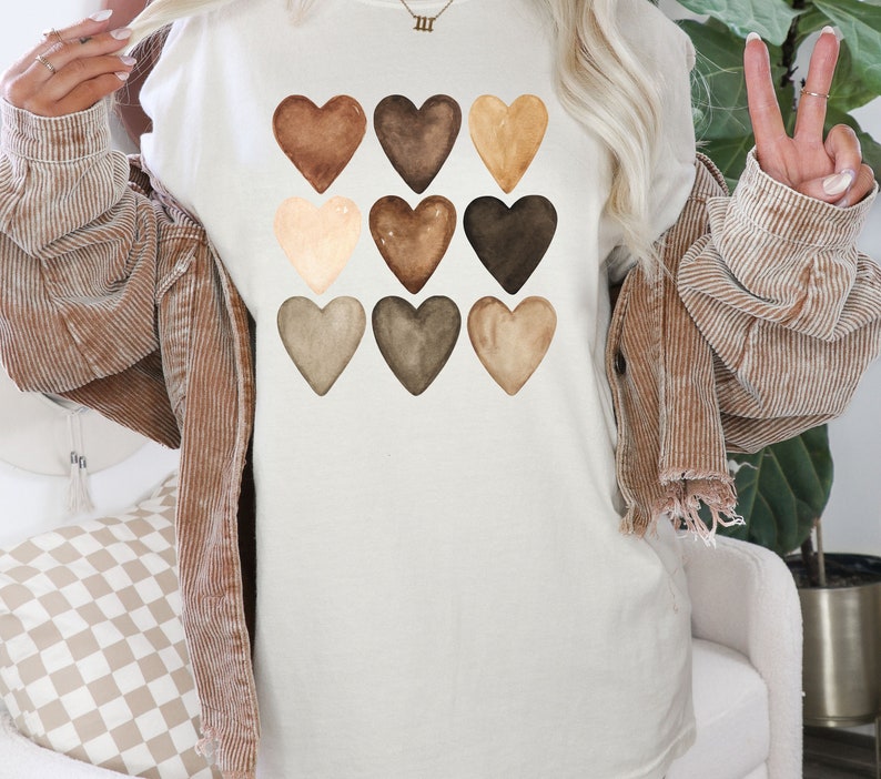 以下が含まれることがあります： クリーム色のTシャツ。9つの水彩画のハートが様々な茶色で描かれています。ハートはグリッドパターンに配置され、シンプルながらもインパクトのあるデザインです。柔らかい素材で作られており、カジュアルウェアに最適です。