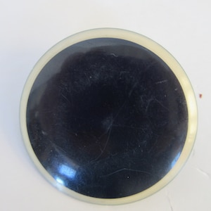 May include: A round, black button with a white rim. The button has a smooth, glossy finish.