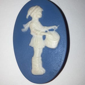 May include: Oval blue cameo featuring a white relief of a figure playing a drum. The figure is wearing a hat, scarf, and tunic. The cameo is a classic design, likely made of ceramic or porcelain.