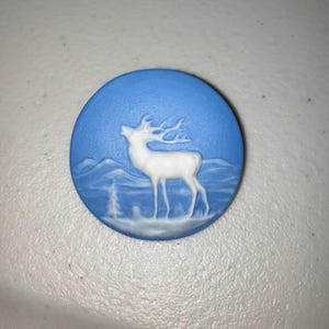 May include: A round, blue decorative piece with a white relief of a deer in a mountainous landscape. The deer is standing with its head raised, and the background features mountains and trees.