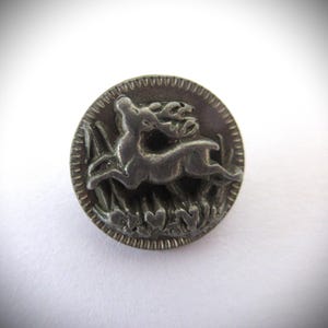 May include: A round, silver-toned button with a detailed relief of a deer leaping through tall grass. The button has a textured edge and is set against a white background. The deer is the central focus of the design.