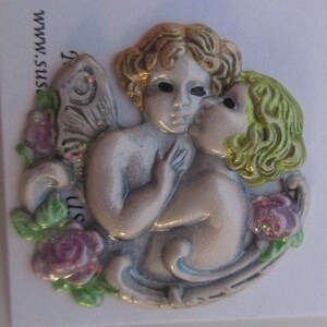 May include: A white ceramic figurine of two angels kissing. The angels have wings and are surrounded by pink and green flowers. The figurine is decorated with glitter.