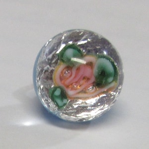 May include: A glass bead with a silver foil background and a pink, green, and white swirl design.