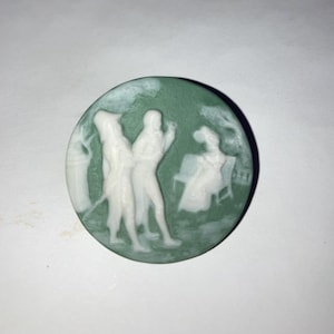 May include: A round, green cameo with a white relief design depicting figures in a park setting. The cameo features a detailed scene of people, trees, and a bench. The cameo is likely made of ceramic or a similar material.
