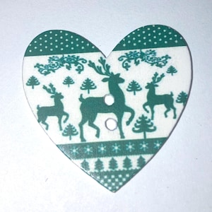 May include: A heart-shaped button featuring a white background with a green pattern of reindeer, pine trees, and snowflakes. The design includes two central holes for attachment, with horizontal bands of decorative motifs across the surface.
