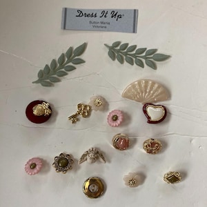 May include: Assortment of vintage-style buttons in various colors and designs, including a heart, a fan, a key, a flower, and a pineapple. The buttons are arranged on a white surface with a green leaf sprig and a card that reads "Dress It Up" Button Mania Victorian.
