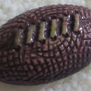 May include: A purple and white ceramic football with a textured surface. The football has the word "NFL" written on it.