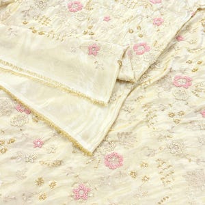 May include: Cream-coloured fabric with detailed floral embroidery and beadwork. The design includes pink and white beaded flowers, with gold and silver accents. The fabric is folded, showing a layered texture and a delicate, shimmering appearance.