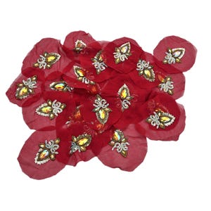 Puede incluir: Un conjunto de pétalos de tela rojos, cada uno adornado con un adorno de tonos joya. Los adornos presentan una gema central en forma de lágrima rodeada de abalorios plateados y dorados, creando un efecto brillante.