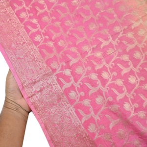 Könnte beinhalten: Ein leuchtend rosa Stoff mit einem sich wiederholenden metallisch goldenen Blumenrankenmuster auf der Oberfläche. Das Textil verfügt über einen breiten, verzierten Rand mit komplizierten gewebten botanischen Motiven und geometrischen Details.